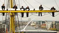 Sechs Männer in blauer Uniform stehen in luftiger Höhe in den Rahen eines Segelmastes. Im Hintergrund die Stadt Dublin.