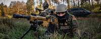 Ein EGB-Soldat liegt mit Maschienengewehr im Anschlag im Gelände, im Hintergrund ein Hubschrauber am Boden
