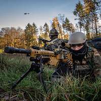 Ein EGB-Soldat liegt mit Maschienengewehr im Anschlag im Gelände, im Hintergrund ein Hubschrauber am Boden