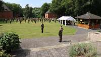 Ein Soldat macht einem anderen Meldung. Im Hintergrund steht eine angetretene Formation.