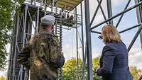 Ein Soldat und eine Zivilistin stehen vor einem großen Metallgerüst, auf dem und unter dem Soldaten stehen.