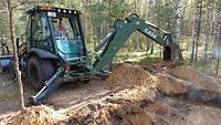 Ein Soldat sitzt in einem Bagger der zwischen Bäumen steht und gräbt ein tiefes Loch in den Waldboden.