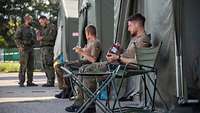 Zwei Soldaten sitzen vor einem olivgrünen Zelt und essen. Im Hintergrund stehen zwei weitere und unterhalten sich.