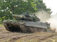 Ein Panzer fährt durch sandiges Gelände, Staub wirbelt auf.