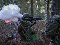 Ein Soldat feuert im Wald kniend eine Panzerfaust ab. Eine Rauchwolke steigt auf.