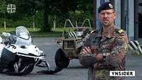Ein Soldat steht vor zwei Fahrzeugen und spricht in die Kamera