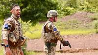 Ein Soldat mit offenem Mund steht hinter einem anderen Soldaten, dieser hat eine Pistole in der Hand und schaut nach vorne 