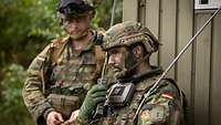 Warten auf den nächsten Befehl Ein Soldat hält sich ein Funkgerät an das Ohr, im Hintergrund steht ein weiterer und sieht ihn an.