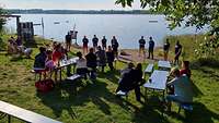 Mehrere Menschen sitzen an zwei langen Biertischen am Ufer eines Sees. Vor ihnen stehen Tauchlehrer.