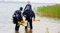 Zwei Menschen stehen mit Tauchausrüstung im Wasser. Ein anderer beobachtet sie.