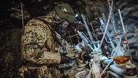 Ein Soldat mit ABC-Schutzmaske steht mit einem Gerät in Hand vor Glaskolben und anderem Labormaterial in einer Höhle