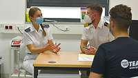 Patienten ansehen und beobachten Eine Soldatin und ein Soldat in weißer Arztkleidung sitzen hinter einem Tisch, davor ein Patient in ziviler Kleidung