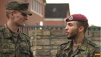 Zwei Soldaten stehen in Uniform vor einer Kasernenmauer und sprechen miteinander. 