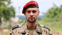 Ein Soldat mit rotem Barett steht vor einem grünen Hintergrund und schaut in die Kamera 