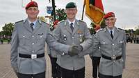 Drei Soldaten in Dienstanzug stehen nebeneinander vor einer Flagge auf einem Platz vor einer Formation von Soldaten.