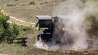 Ein Brückenpanzer fährt durch das Gelände, Staub wirbelt auf.