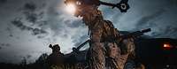 Ein Soldat kniet mit Waffe über einer am Boden liegenden Person bei Dunkelheit, dahinter ein Hubschrauber am Himmel