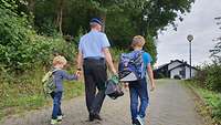 Rückenansicht von einem Soldaten und zwei Kindern mit Schulranzen, die händschenhaltend über einen Weg gehen.