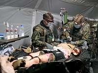 Zwei Soldaten stehen an einem Feldoperationstisch in einem Sanitätszelt und versorgen eine liegende Übungspuppe 