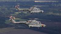 Zwei graue Hubschrauber fliegen nebeneinander über Land.