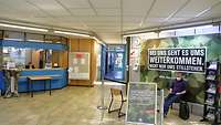  Foyer mit einer Person sitzend an der Anmeldung. Eine weitere Person wartet mit Rucksack und einer Mappe in der Hand.
