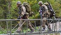 Zwei Soldaten überqueren einen breiten Bach über eine schmale, provisorische Metallbrücke.