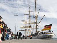 Einlaufen „Gorch Fock“ 2er-Bilderpanel 2 Mehrere Menschen blicken auf ein weißes Segelschulschiff der Deutschen Marine.