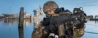 Ein Soldat mit Waffe im Anschlag, dahinter Wasser und graue Schiffe im Hafen