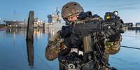 Spezialkräfte Ein Soldat mit Waffe im Anschlag, dahinter Wasser und graue Schiffe im Hafen