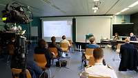 Eine Gruppe Menschen sitzt im Hörsaal und hört einer Frau zu, die über WebEx eine Präsentation hält.