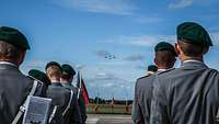 Überflug Eine Formation von Soldaten steht mit Blickrichtung zum Hintergrund des Bildes. Ihnen kommen am Himmel vier Flugzeuge entgegen.