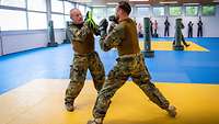 Zwei Soldaten boxen in einer hellen Halle auf gelb-blauem Boden.