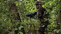 Ein Soldat in Gefechtsanzug und blauer Übungswaffe läuft durch den dicht bewachsenen Wald.