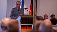 Ein Soldat in Dienstanzug steht hinter einem Rednerpult und spricht, am Rednerpult und neben ihm die deutsche Nationalflagge.