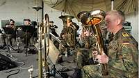 Mehrere Soldaten spielen sitzend unter einem Dach an ihren goldglänzenden Blasinstrumenten.