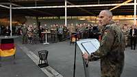 Soldaten und zivile Gäste stehen draußen unter einer Überdachung und hören zu, während ein Soldat eine Rede am Pult hält.