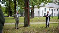 Ein großes Holzkreuz lehnt an einem Baum. Daneben stehen zwei Pfarrer
