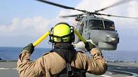 Ein Soldat mit neongelben Helm gibt auf dem Flugdeck der „Bonn“ das Sichtzeichen zum Start mit zwei gelben Lichtern. 