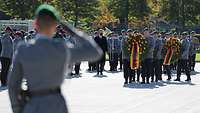 Im Vordergrund ein Soldat, der militärisch grüßt. Im Hintergrund Soldaten bei der Kranzniederlegung.