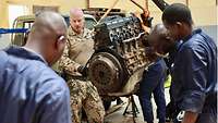 Malische Männer stehen mit einem deutschen Soldaten um einen an einem Kran hängenden Motorblock