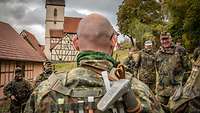 Soldaten stehen bei einer Ausbildung im Halbkreis auf einer Wiese neben historischen Gebäuden.