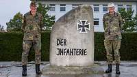 Zwei Soldaten in Uniform stehen neben einem großen grauen Stein mit der Inschrift „Der Infanterie“.
