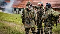 Mehrere Soldaten stehen in einem Schießtraining neben einem Gebäude auf einer grünen Wiese.
