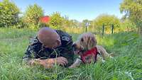 Ein Soldat und ein Hund liegen auf dem Bauch im hohen Gras und schauen sich an.