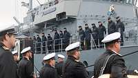 Mehrere Menschen in blauer Uniform stehen an Oberdeck eines grauen Schiffes. Davor Menschen in schwarzer Uniform.