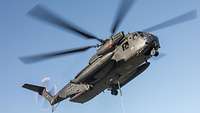Ein fliegender Hubschrauber vom Typ CH-53. An seiner Unterseite ist der Lasthaken mit einer textilen Kette zu sehen.