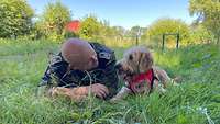 Soldat und Therapiehund liegen auf dem Bauch im hohen Gras und schauen sich an