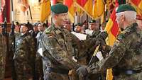 Ein Soldat überreicht einem anderen die Truppenfahne, im Hintergrund stehen Soldaten mit zahlreichen Truppenfahnen.