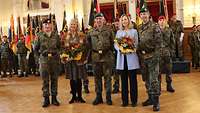 Drei Soldaten und zwei Zivilistinnen stehen nebeneinander. Die Frauen halten Blumensträuße in den Händen.