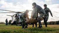 Im Vordergrund tragen mehrere Soldaten eine Person. Im Hintergrund sind weitere Personen und ein Hubschrauber